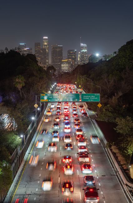 Busy Los Angeles freeway traffic at night with downtown skyline in the background, captured from above.